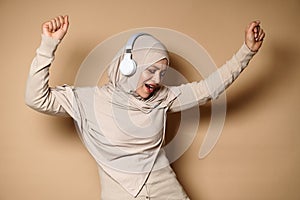 Happy Muslim woman in hijab and headphones cute smiles and relaxes, dances while listening to the music against beige background.