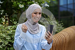 Happy Muslim woman in hijab surprised by online win, sitting on street holding phone in hand, lucky lottery