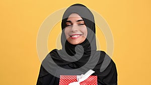 Happy Muslim woman holding red gift box, looking inside and surprised, portrait in orange background