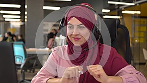 A happy Muslim woman wearing a hijab and headphones listens to music and dances in the workplace