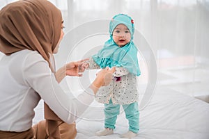 Happy muslim young mother and adorable little baby daughter in hijab on white bed in bedroom at home. Muslim mom hands holding