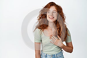 Happy natural girl with red hair and freckles, holding hand on heart, smiling and looking asie with cheerful face photo