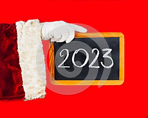 Happy New Year 2023- holiday greeting. Santa Claus`s hand is holding a blackboard with the new year 2023 number written in chalk.