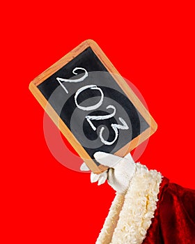 Happy New Year 2023- holiday greeting. Santa Claus`s hand is holding a blackboard with the new year 2023 number written in chalk.