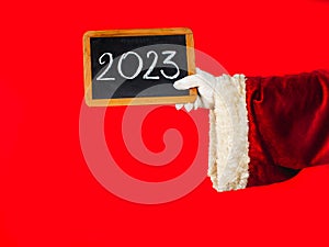 Happy New Year 2023- holiday greeting. Santa Claus`s hand is holding a blackboard with the new year 2023 number written in chalk.