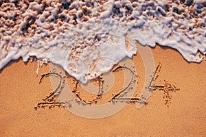 Happy New Year 2024 text written on the beach sand and ocean wave washing the sea shore