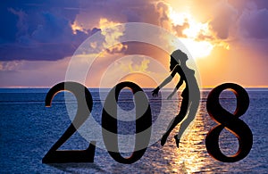 Happy new year card 2018. Silhouette of young woman on the beach jumping as a part of the Number 2018 sign with sunset background