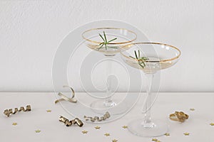 Happy New Year composition. Two champagne wine glasses with golden rim and green rosemary. White table, golden confetti