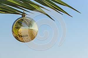 Happy New Year and Merry Christmas on beach concept. Golden Christmas ball on palm branch against blue sky. Copy space