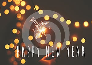 Happy New Year text sign on glowing sparkler in hand on background of golden christmas tree lights in dark festive room. Fireworks