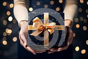 Happy New Year! A woman hands present an elegant present gift box with golden ribbon , background with gold bokeh, top view