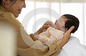 Happy newborn baby girl or boy sleeping on mother arm. Smiling mother touching chin of infant daughter or son on bed at home.