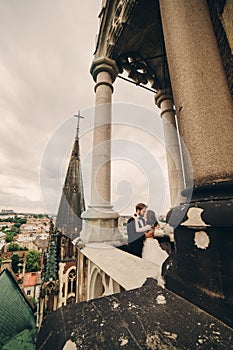 Happy newlywed. beautiful bride and stylish groom are kissing on the balcony of old gothic cathedral with panoramic city views