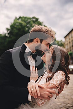 Happy newlywed couple hugging and kissing in old European town street, gorgeous bride in white wedding dress together with