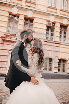 Happy newlywed couple hugging and kissing in old European town street, gorgeous bride in white wedding dress together with
