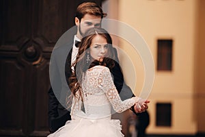 Happy newlywed couple hugging and kissing in old European town street, gorgeous bride in white wedding dress together with