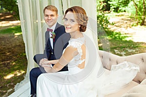 Happy newlyweds holding hands and sitting on beige sofa in park