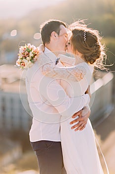 Happy newlyweds kissing while bride holds bouquet on cityscape background