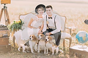 Happy newlyweds are sitting with the three little dogs on the old-fashioned sofa surrouned by the vintage suitcases