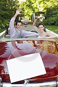 Happy newlyweds waving in convertible with blank sign in foreground