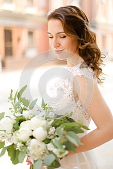 Happy nice bride keeping bouquet of flowers and wearing white dress.