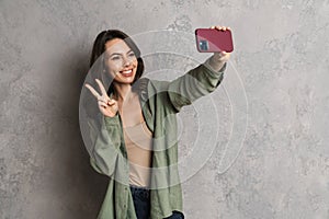 Happy nice girl gesturing peace sign while taking selfie on cellphone