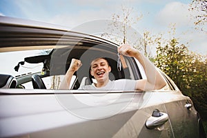 Happy Novice driver keeps fist up tight looking out of the driver window of his new car holding key from a new car. Successful