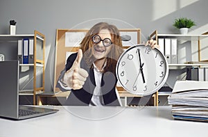 Happy office worker sitting at desk, holding clock that says it's 5 pm and giving thumbs-up