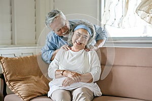 happy old asian husband take care and massage shoulder elderly wife sitting on sofa in living room at home . Lovely asia senior