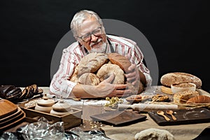 Happy old baker looking at camera and smiling while hugging loaves of bread