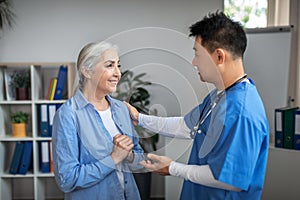 Happy old caucasian female patient at meeting thanks to millennial japanese doctor in uniform in office