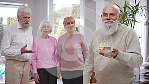 Happy old Caucasian man with piece of birthday cake looking back at friends singing clapping and looking at camera