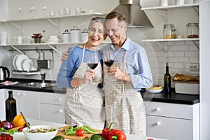 Happy old couple drinking wine preparing vegetable salad in kitchen at home.