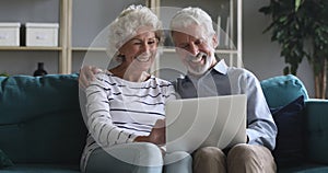 Happy old couple talking laughing using laptop sit on sofa