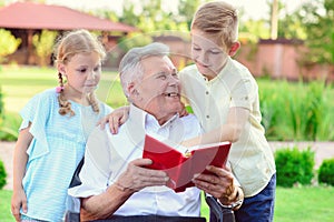 Happy old grandfather reading book for cute children in garden