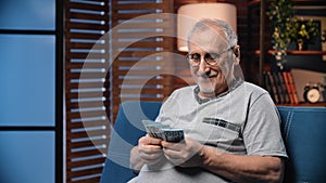 Happy old man counting money at home in evening, portrait of senior person with dollars banknotes