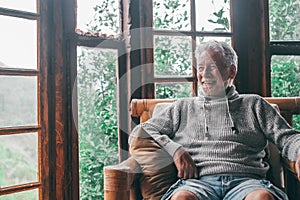 Happy old man spending leisure time at home looking out through window. Smiling senior man with gray hair and sweater relaxing on