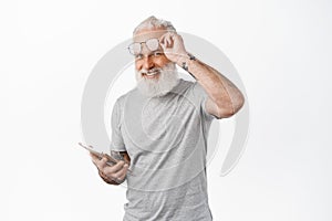 Happy old man with tattoos, take off glasses and using digital tablet, smiling at camera, standing in grey t-shirt