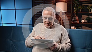 Happy old man viewing pictures and chatting online in social media by tablet, male portrait in home