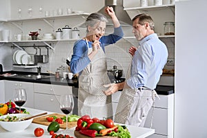 Happy old senior 50s couple having fun dancing cooking together in kitchen.