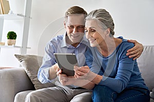 Happy old senior grandparents using digital tablet sitting on couch at home.