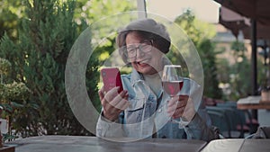 Happy old woman having a video call with friends during sitting in restaurant and drinking red wine on vacation