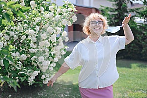 Happy old woman takes off the mask and sniffs a blossoming apple tree. A female pensioner enjoys fresh air on a warm