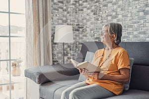 Happy older blonde woman sitting on comfortable sofa in living room, reading interesting book. Smiling elder retired grandmother