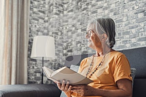 Happy older blonde woman sitting on comfortable sofa in living room, reading interesting book. Smiling elder retired grandmother