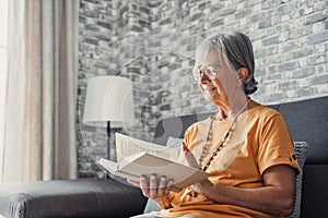 Happy older blonde woman sitting on comfortable sofa in living room, reading interesting book. Smiling elder retired grandmother