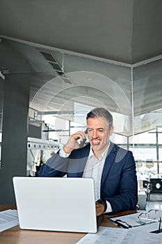 Happy older business man talking on cell phone using laptop in office.