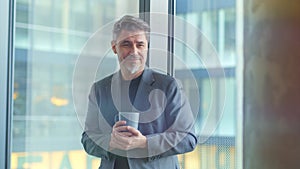 Happy older businessman taking break in office drinking coffee standing in window and daydreaming.