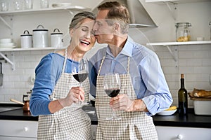 Happy older couple hugging, drinking wine in kitchen celebrating Valentines Day.
