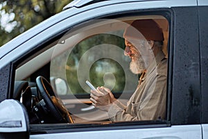 Happy older man traveler sitting in camper van using mobile in camping.